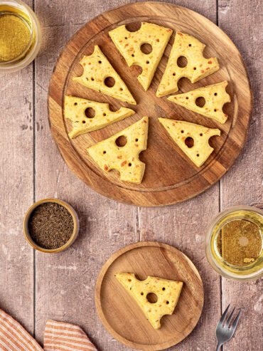 Käse-Cracker in Form von Emmentaler-Dreiecken mit ausgestochenen Löchern auf einem runden Holzbrett, daneben Kümmel und ein Glas Weißwein