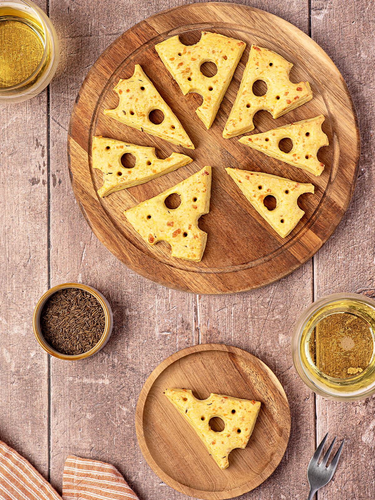 Käse-Cracker in Form von Emmentaler-Dreiecken mit ausgestochenen Löchern auf einem runden Holzbrett, daneben Kümmel und ein Glas Weißwein