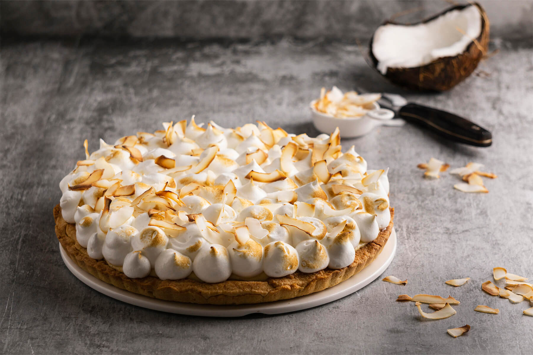 Ganze Kokos-Sahne-Tarte auf hellem Brett mit halbierter Kokosnuss und gerösteten Kokosflocken