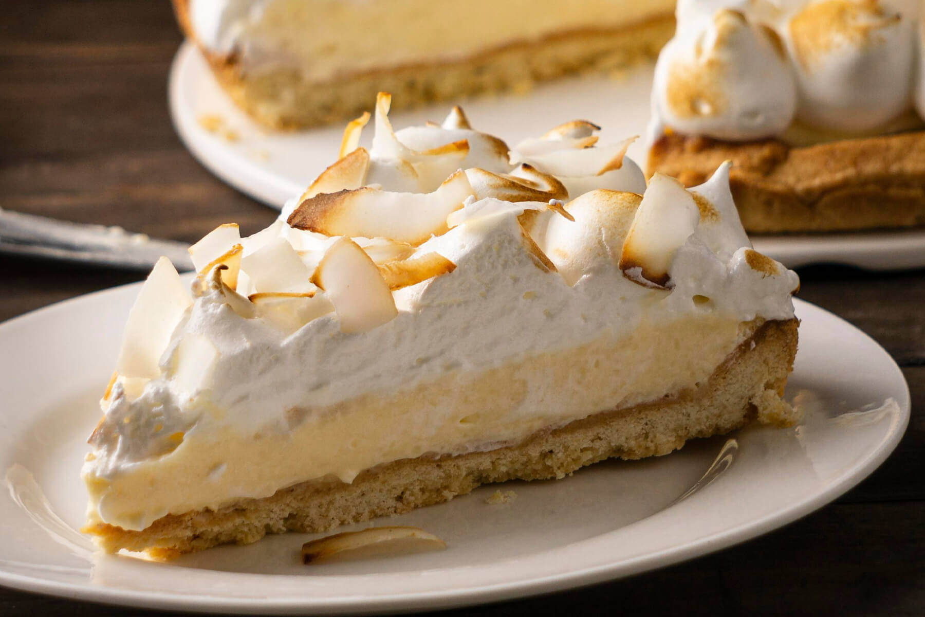 Stück Kokos-Sahne-Tarte im Querschnitt mit Mürbeteigboden, dicker Kokoscreme und gebackenem Baiser mit gerösteten Kokosflocken