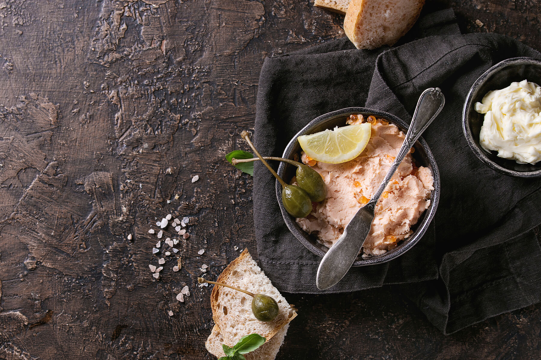 Lachs-Pâté in dunkler Schale mit Kapern-Beeren und Zitrone, daneben Butter, Bauernbrot und Meersalz auf dunklem Stein