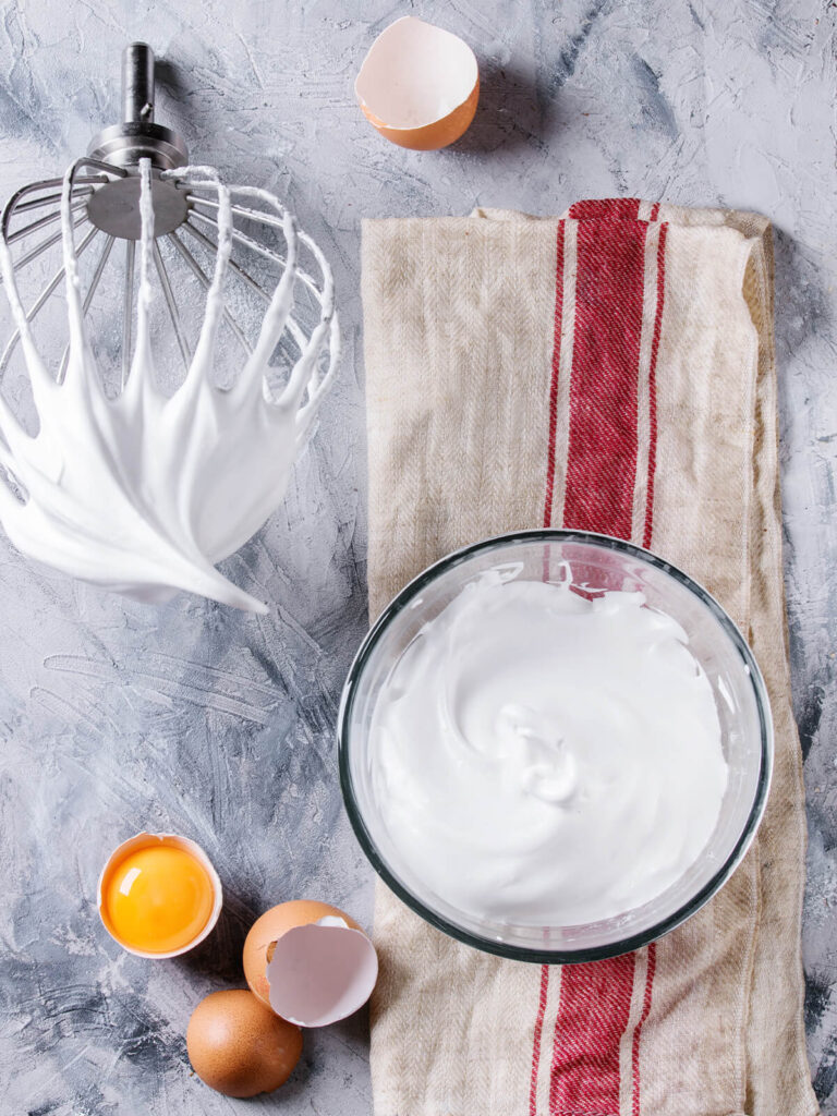Fertig aufgeschlagene weiße Meringue-Masse in einer Glasschüssel neben Schneebesen und getrenntem Eigelb