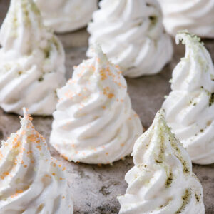 Nahaufnahme weißer Meringue-Tannenbäume mit orangefarbenen und hellgrünen Zuckerkristallen auf einem dunklen Backblech