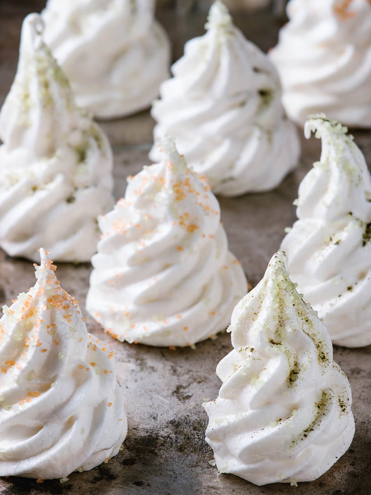 Nahaufnahme weißer Meringue-Tannenbäume mit orangefarbenen und hellgrünen Zuckerkristallen auf einem dunklen Backblech