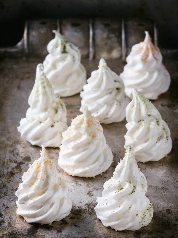 Weiße Meringue-Tannenbäume mit orangefarbenen und hellgrünen Zuckerkristallen auf einem dunklen Backblech