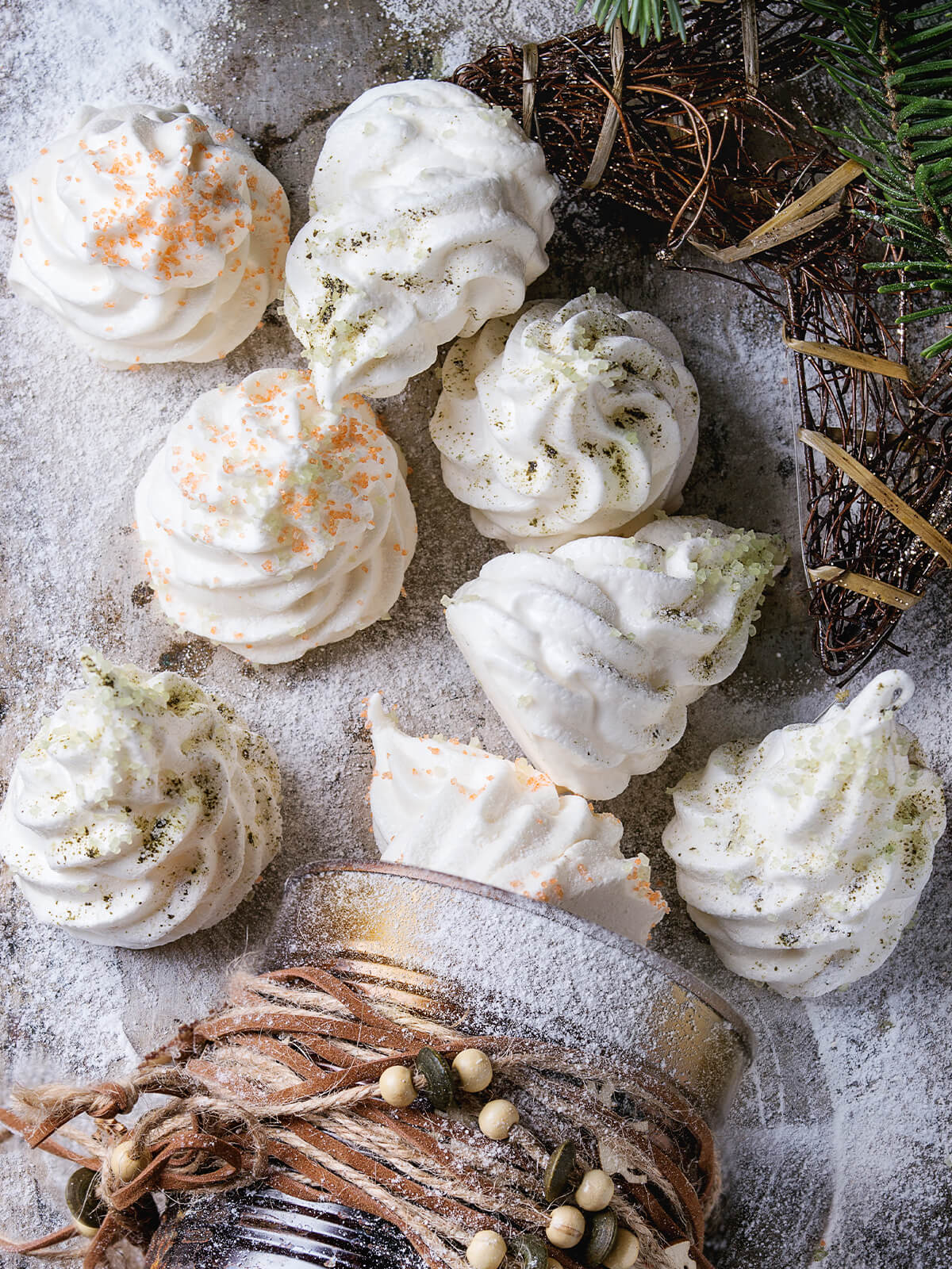 Meringue-Tannenbäume mit Zuckerkristallen von oben auf einer mit Puderzucker bestäubten Fläche, daneben Tannenzweig und Weihnachtsdeko
