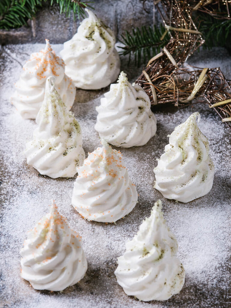 Meringue-Tannenbäume mit bunten Zuckerkristallen auf Puderzucker-Schnee, dahinter ein Tannenzweig und ein Stern aus Zweigen