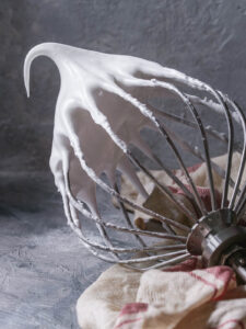 Dichte, glänzende Meringue am waagerecht gehaltenen Schneebesen, die Spitze steht und biegt sich vorne leicht um