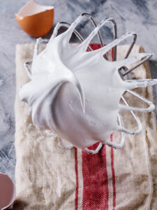 Steif geschlagene, glänzende Meringue am Schneebesen der Küchenmaschine, daneben Eierschalen