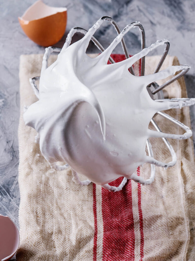 Steif geschlagene, glänzende Meringue am Schneebesen der Küchenmaschine, daneben Eierschalen
