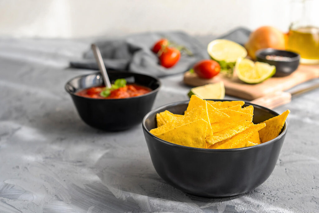 Mexikanischer Tomaten-Dip in dunkler Schale, daneben eine Schale knuspriger Tortilla-Chips mit Limetten und Tomaten