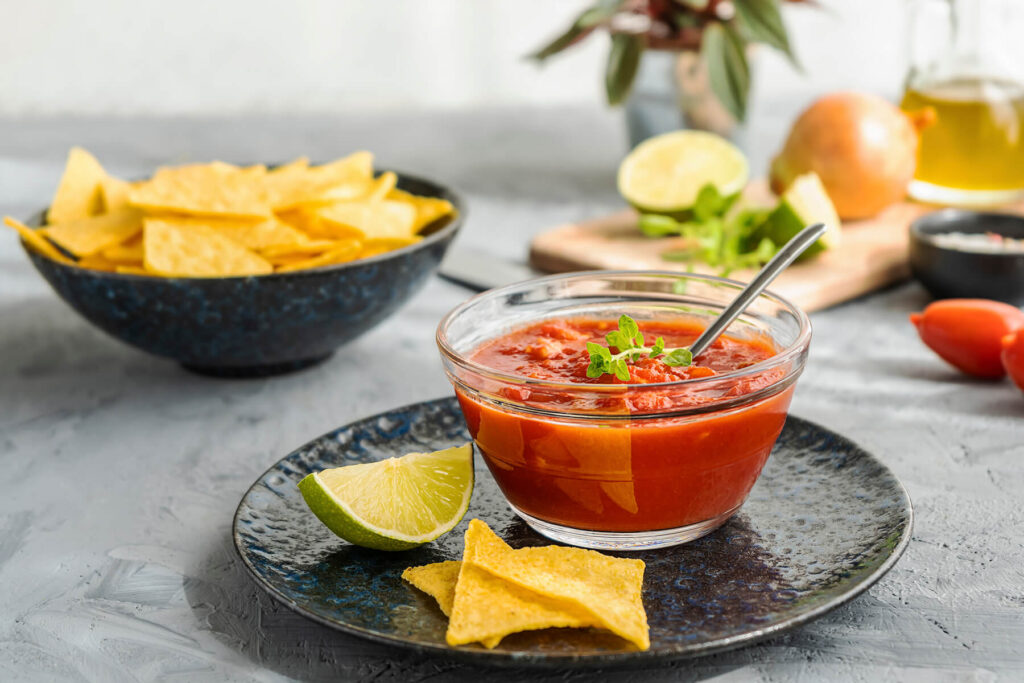 Mexikanischer Tomaten-Dip in einer Glasschale mit Koriander-Zweig, daneben Limettenviertel und Tortilla-Chips