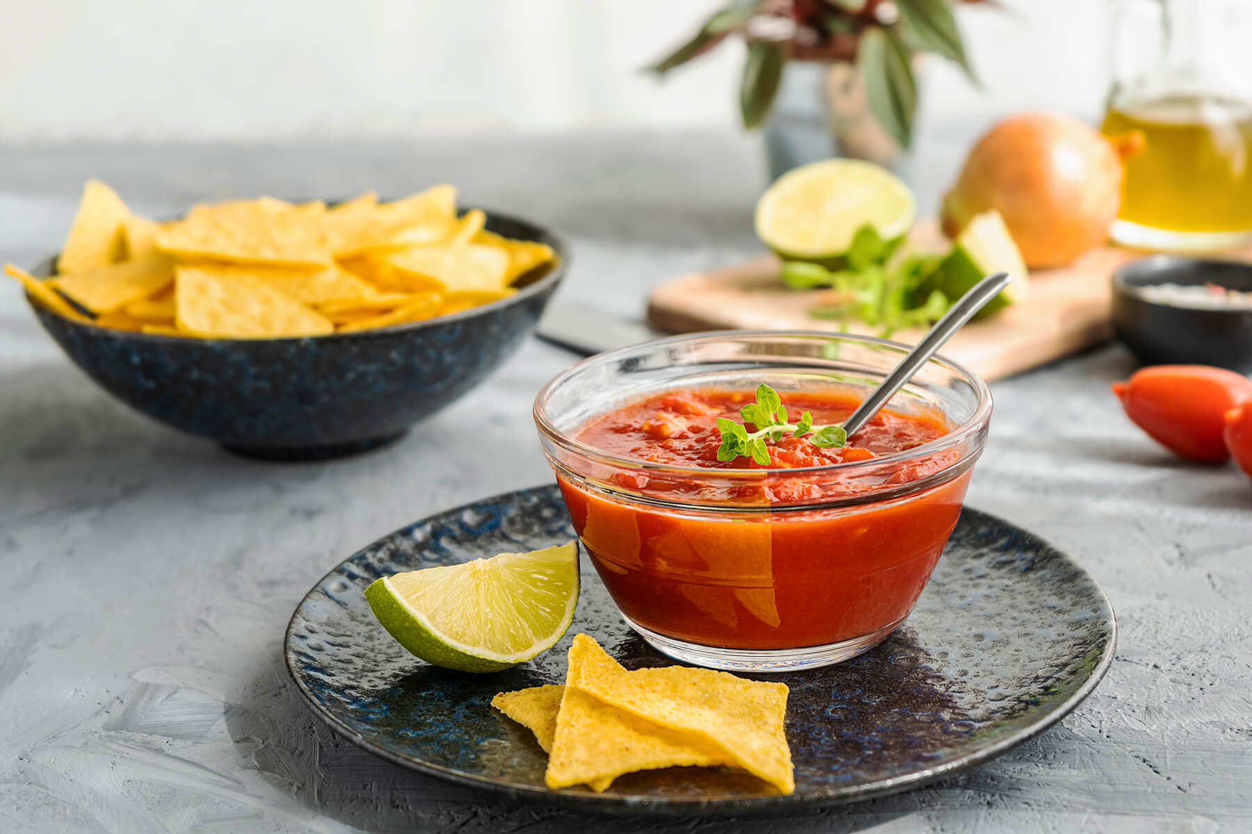 Mexikanischer Tomaten-Dip in einer Glasschale mit Kräuterzweig, daneben Limettenviertel und Tortilla-Chips