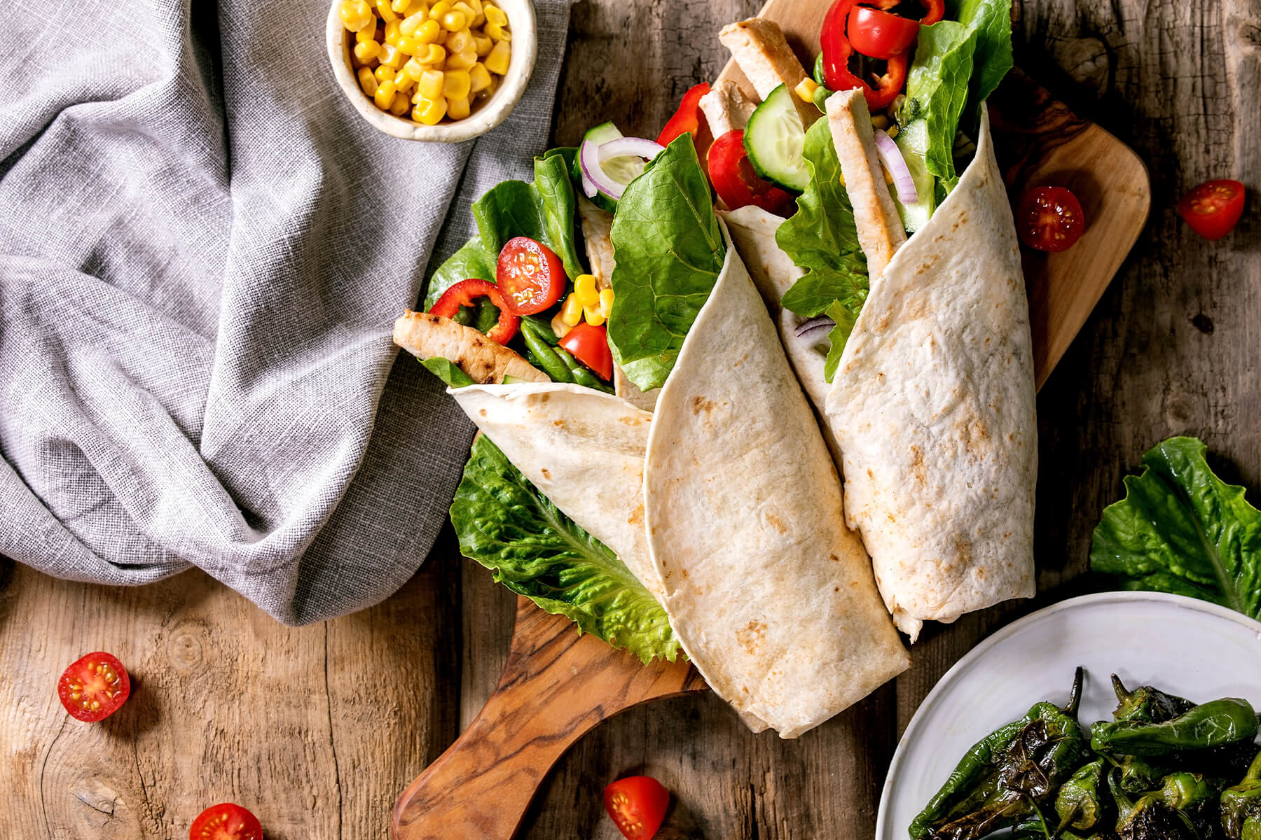 Zwei übergeklappte Wraps auf einem Holzbrett mit Leinentuch, daneben ein Schälchen Mais, Cherry-Tomaten und am Bildrand gebratene grüne Pimientos