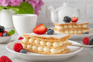 Mille-Feuille mit Vanillecreme, halbierter Erdbeere und Heidelbeere, dahinter ein zweites Stück