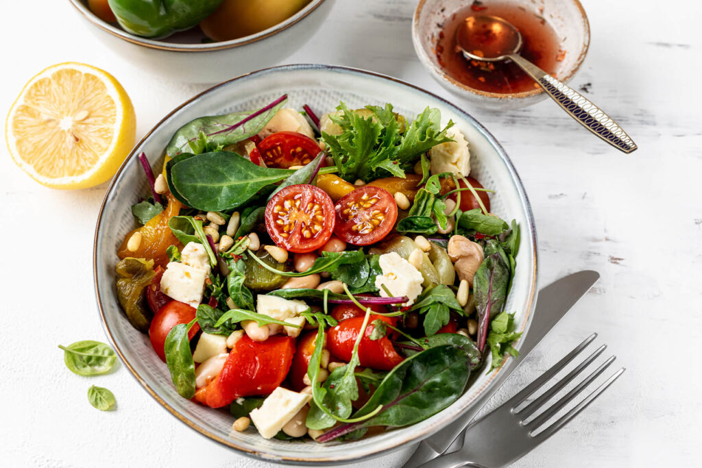 Mediterrane Bowl von oben mit gerösteter Paprika, weißen Bohnen, Feta und gemischten Greens, daneben eine halbe Zitrone und ein Schälchen Zitronen-Olivenöl-Dressing
