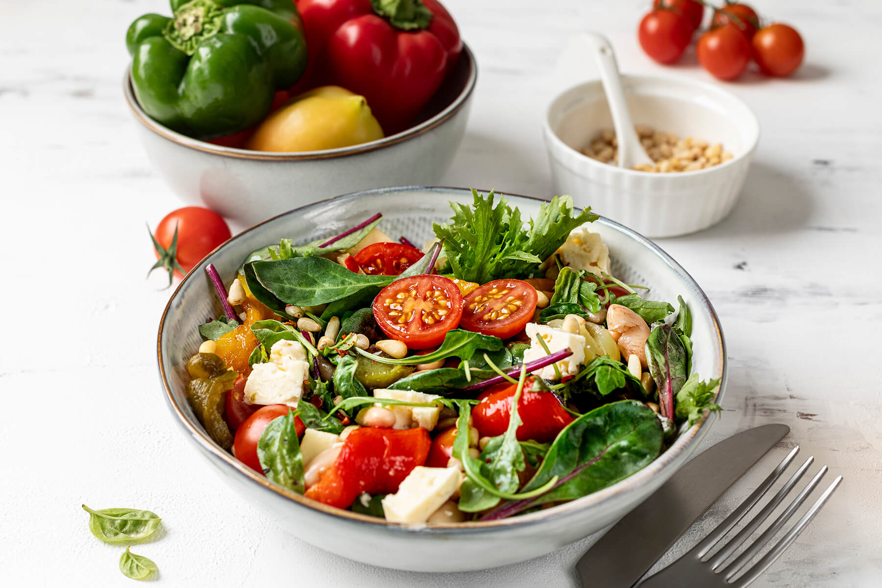 Mediterrane Bowl mit weißen Bohnen, gerösteten Paprika-Stücken in Rot, Gelb und Grün, Feta und gemischten Blattsalaten, im Hintergrund eine Schale mit frischen grünen und roten Paprika