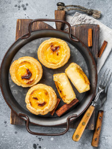 Pastéis de Nata mit Zimtstangen in einer dunklen Servierpfanne, ein Törtchen aufgeschnitten