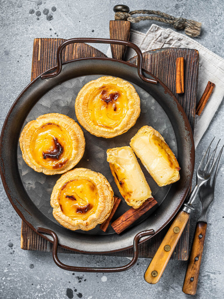 Pastéis de Nata mit Zimtstangen in einer dunklen Servierpfanne, ein Törtchen aufgeschnitten