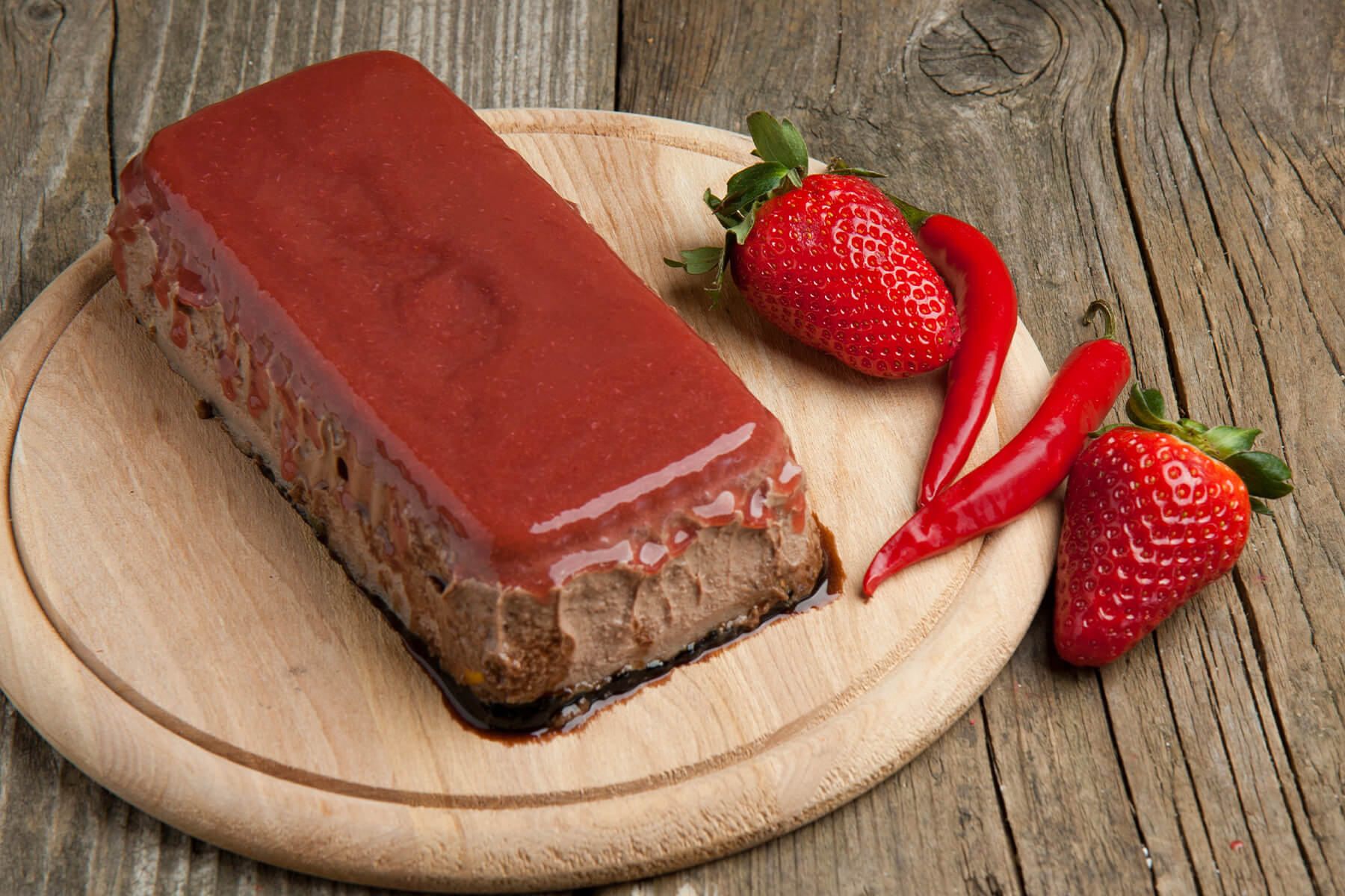 Ganze Leberpastete-Terrine mit glänzender Erdbeer-Chili-Gelee-Decke auf rundem Holzbrett, daneben frische Erdbeeren und rote Chilischoten