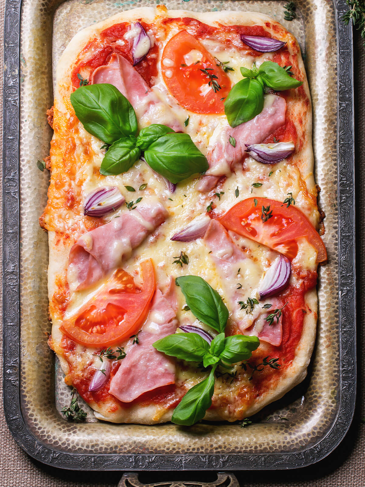 Rechteckige Blechpizza mit Tomatensauce, Käse, Schinken, Tomatenscheiben, roten Zwiebeln und Basilikum
