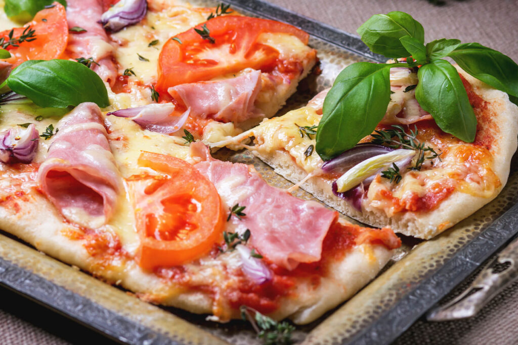 Rechteckige Blechpizza mit Schinken, Tomaten, roten Zwiebeln und Basilikum