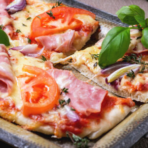 Rechteckige Blechpizza mit Schinken, Tomaten, roten Zwiebeln und Basilikum