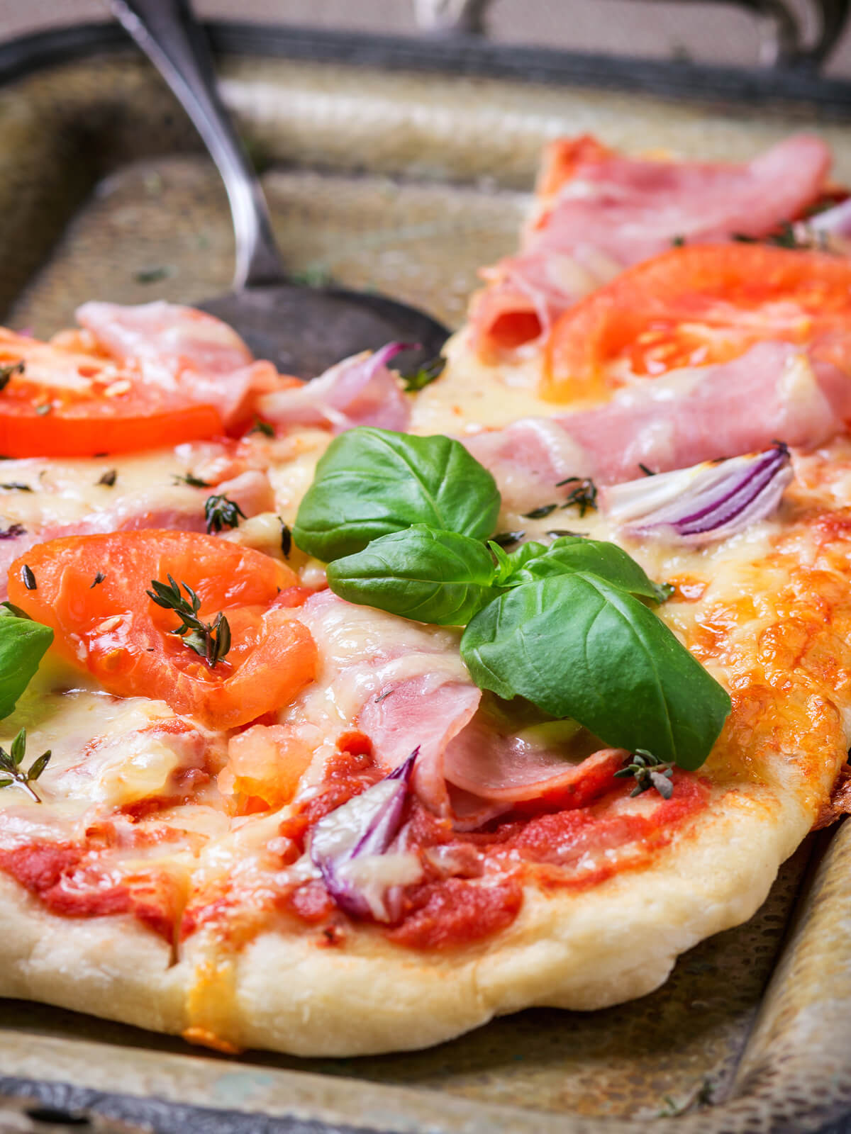 Nahaufnahme der Blechpizza mit Schinken, Tomatenscheiben, roten Zwiebeln, Thymian und Basilikum