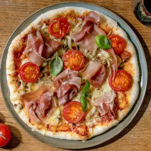 Pizza Napoletana mit Parmaschinken, halbierten Cocktailtomaten, Champignons und Basilikum auf grünem Teller
