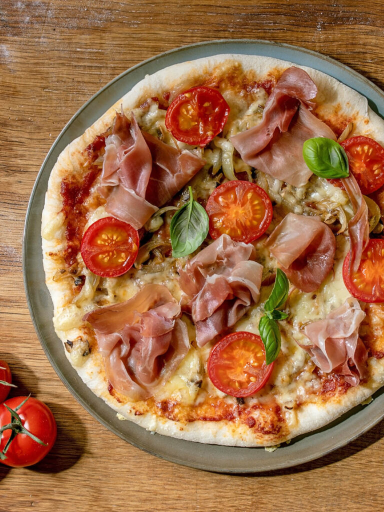 Fertig gebackene Pizza Napoletana mit Parmaschinken, halbierten Cocktailtomaten, Champignons und Basilikum auf grünem Teller