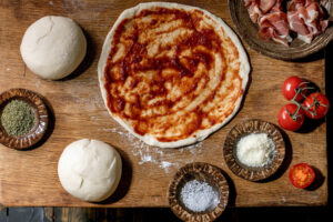 Ausgezogener Pizzateig mit eingekochter Tomatensauce, daneben Teigballen, Parmaschinken, Parmesan, Oregano und Rispentomaten