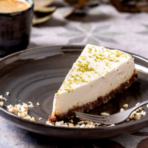 Stück Raw Vegan Cheesecake mit heller Cashew-Füllung auf dunkler Dattel-Mandel-Krume, bestreut mit Limettenschale und gehackten Cashews