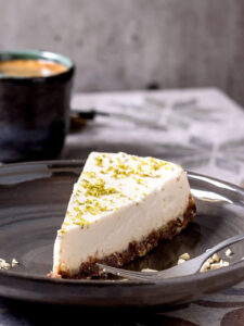 Angeschnittenes Stück Raw Vegan Cheesecake mit Cashew und Limette auf grauem Keramikteller mit Gabel, dahinter eine Tasse Espresso
