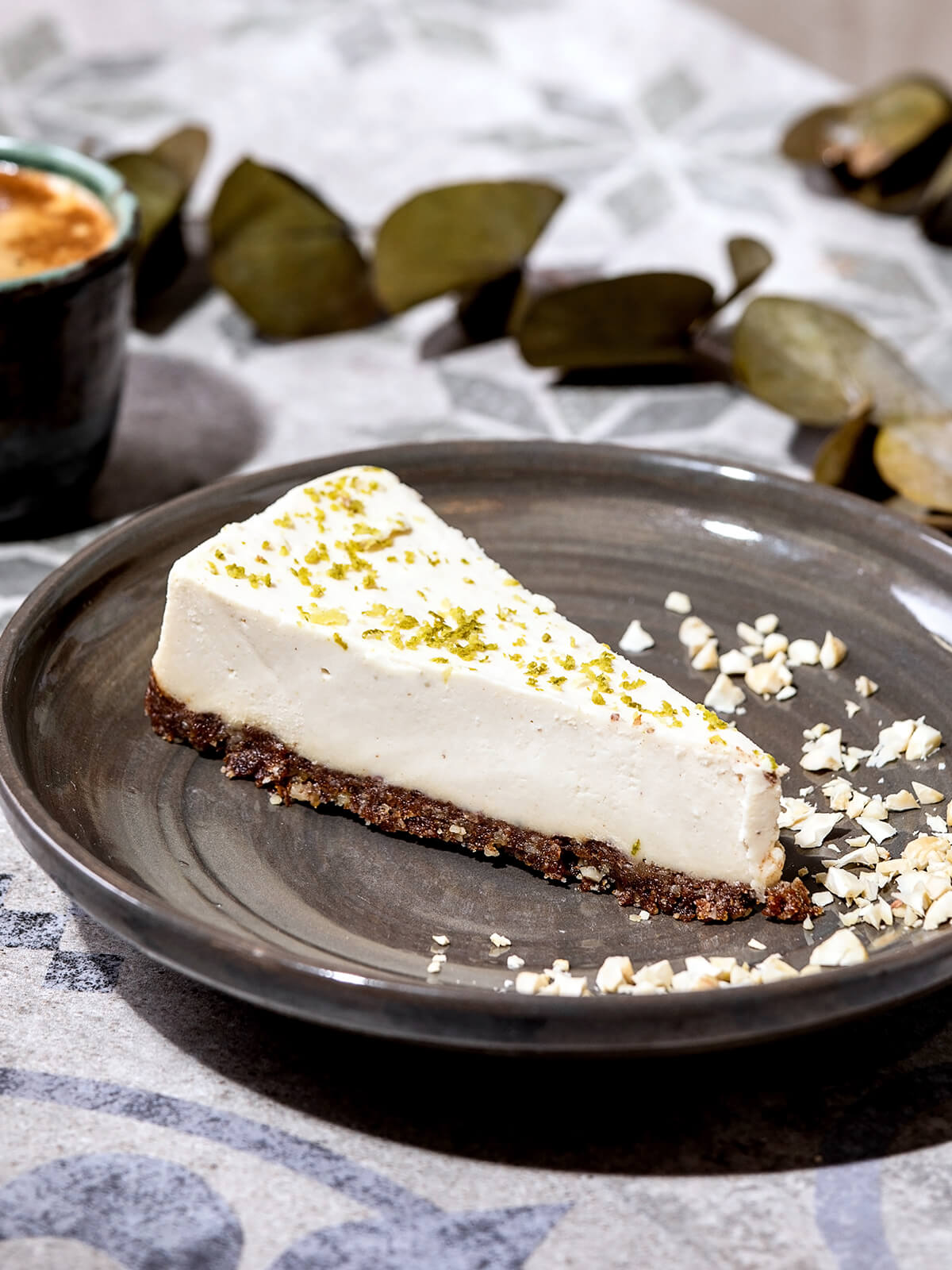 Tortenstück Raw Vegan Cheesecake mit Cashew und Limette auf grauem Keramikteller mit gehackten Cashews und Eukalyptuszweig