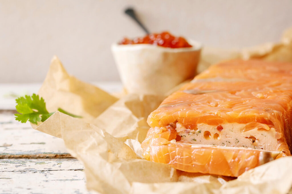Angeschnittene Lachs-Terrine im Profil mit cremiger Frischkäse-Füllung, dahinter eine Schale roter Kaviar