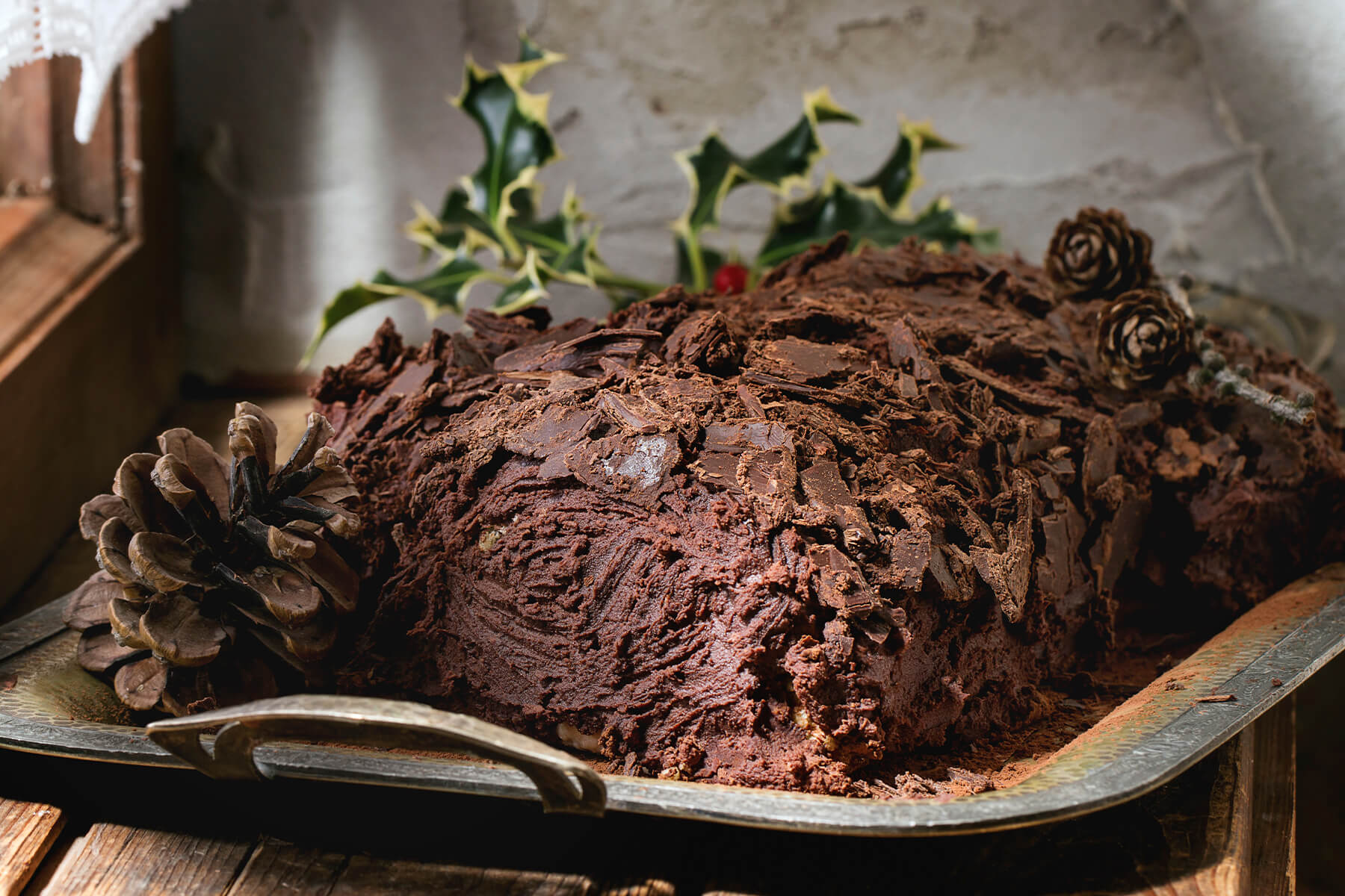 Bûche de Noël aus der Nähe mit grober Rinde aus Schokoladensplittern, Tannenzapfen und Stechpalme