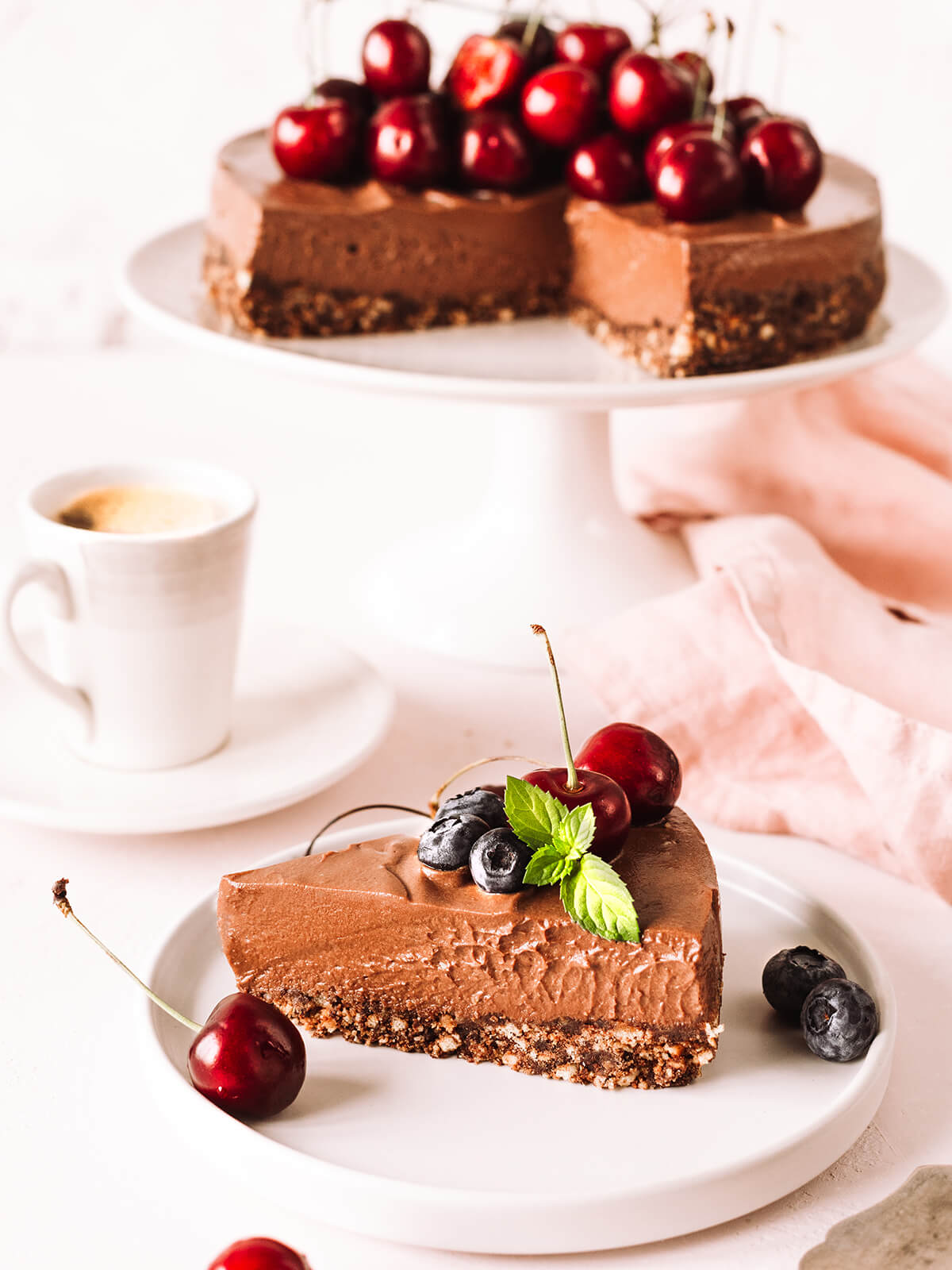 Stück No-Bake Schoko-Cheesecake auf weißem Teller mit Heidelbeeren, Kirsche und Minze, dahinter die angeschnittene Torte
