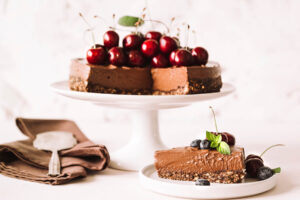 Angeschnittener No-Bake Schoko-Cheesecake mit Kirschen auf Tortenplatte, davor ein Stück auf einem Teller