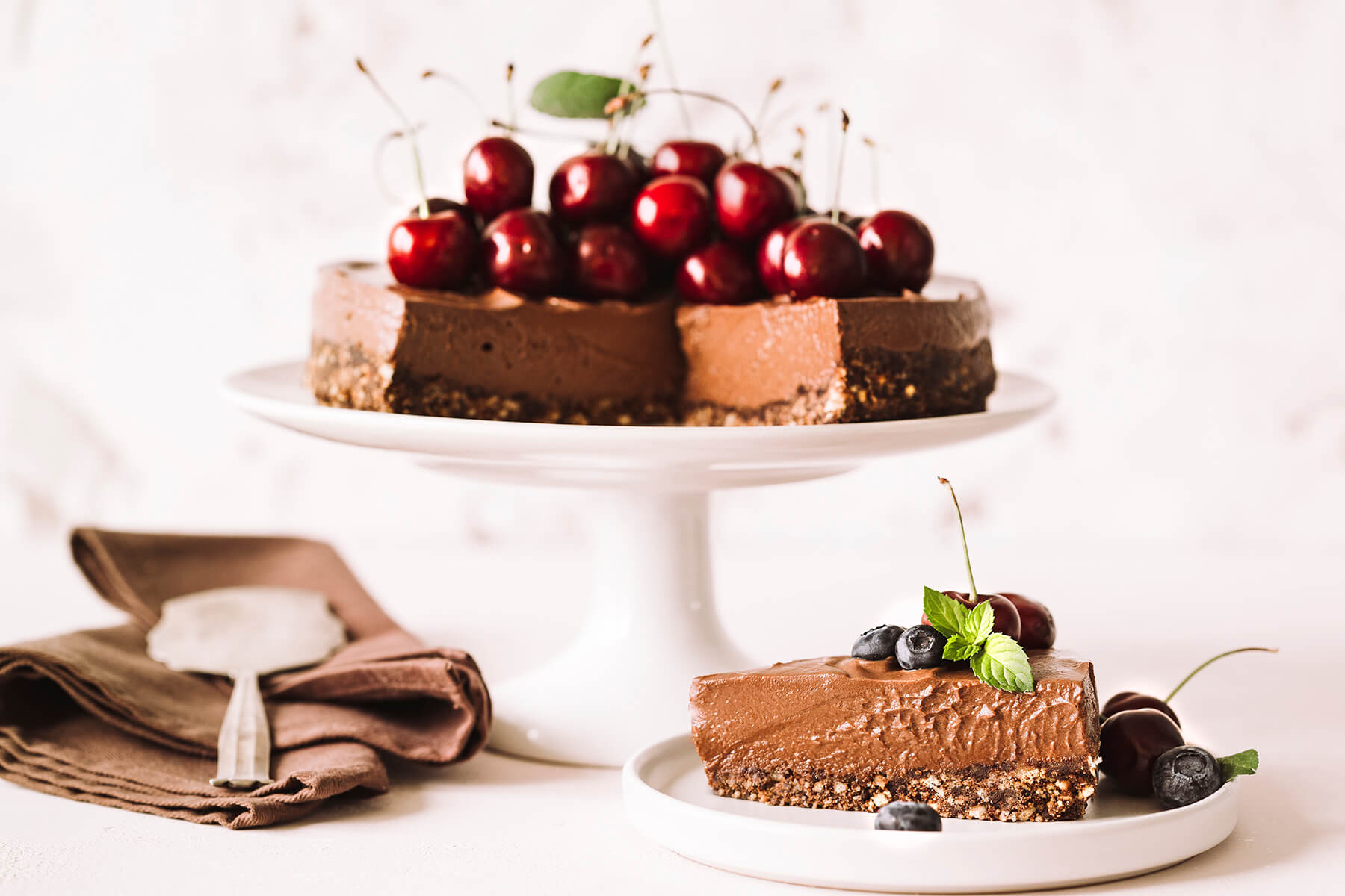 Angeschnittener No-Bake Schoko-Cheesecake mit Kirschen auf Tortenplatte, davor ein Stück auf einem Teller