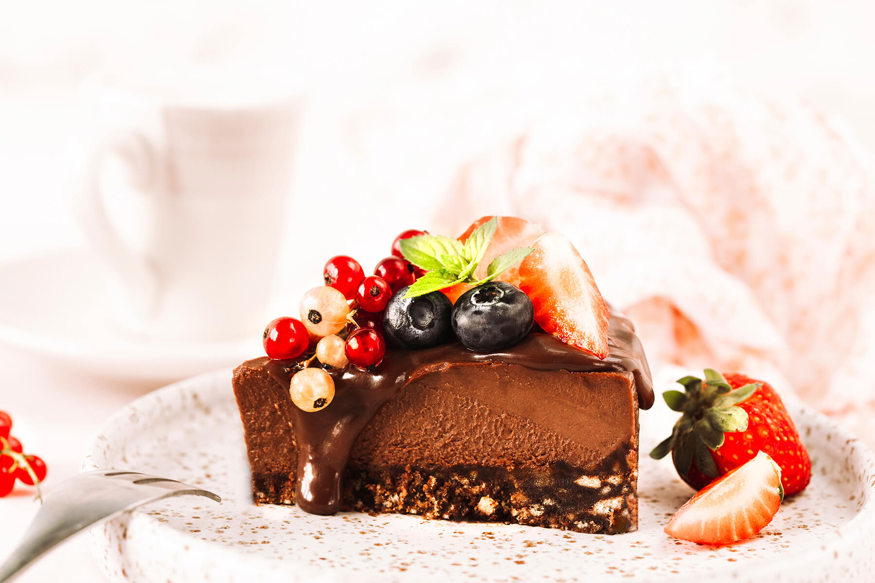 Stück Schoko-Cheesecake mit dunklem Schokoladenguss, Erdbeeren, Johannisbeeren und Heidelbeeren auf gesprenkeltem Teller