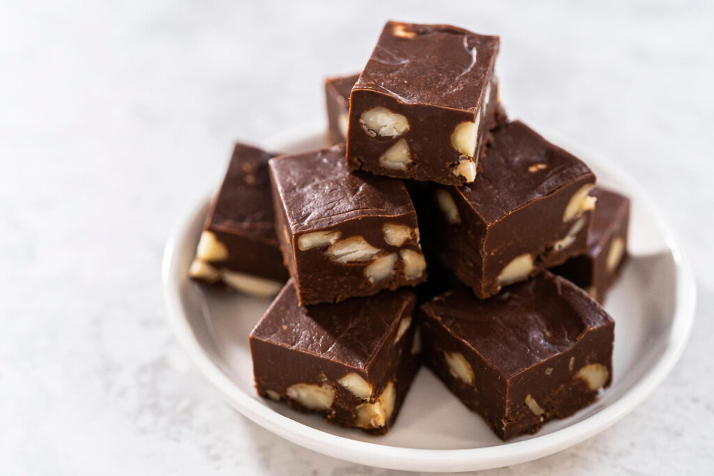 Aufgetürmte Schoko-Macadamia-Fudge-Würfel auf einem hellen Teller mit ganzen Macadamia-Nüssen im Anschnitt