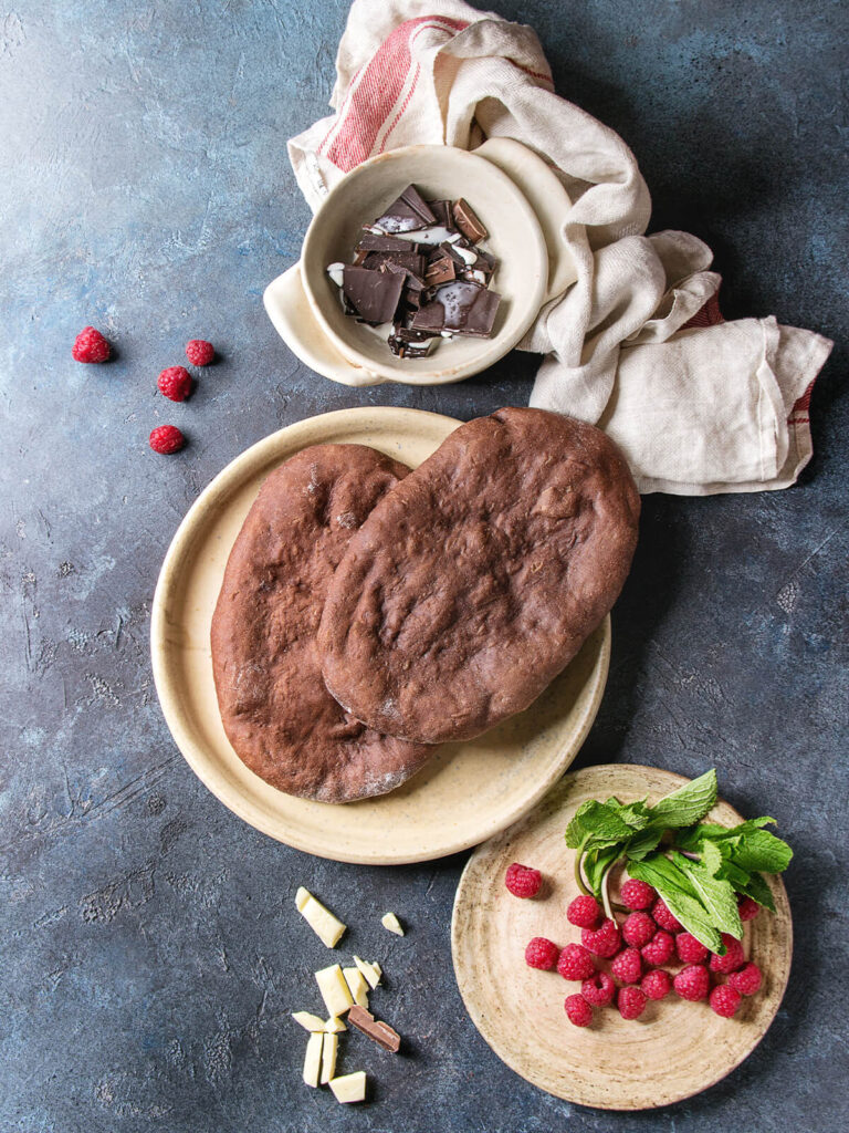Zwei pur gebackene ovale Kakao-Fladen neben Schälchen mit Himbeeren, Schokoladenstücken und frischer Minze