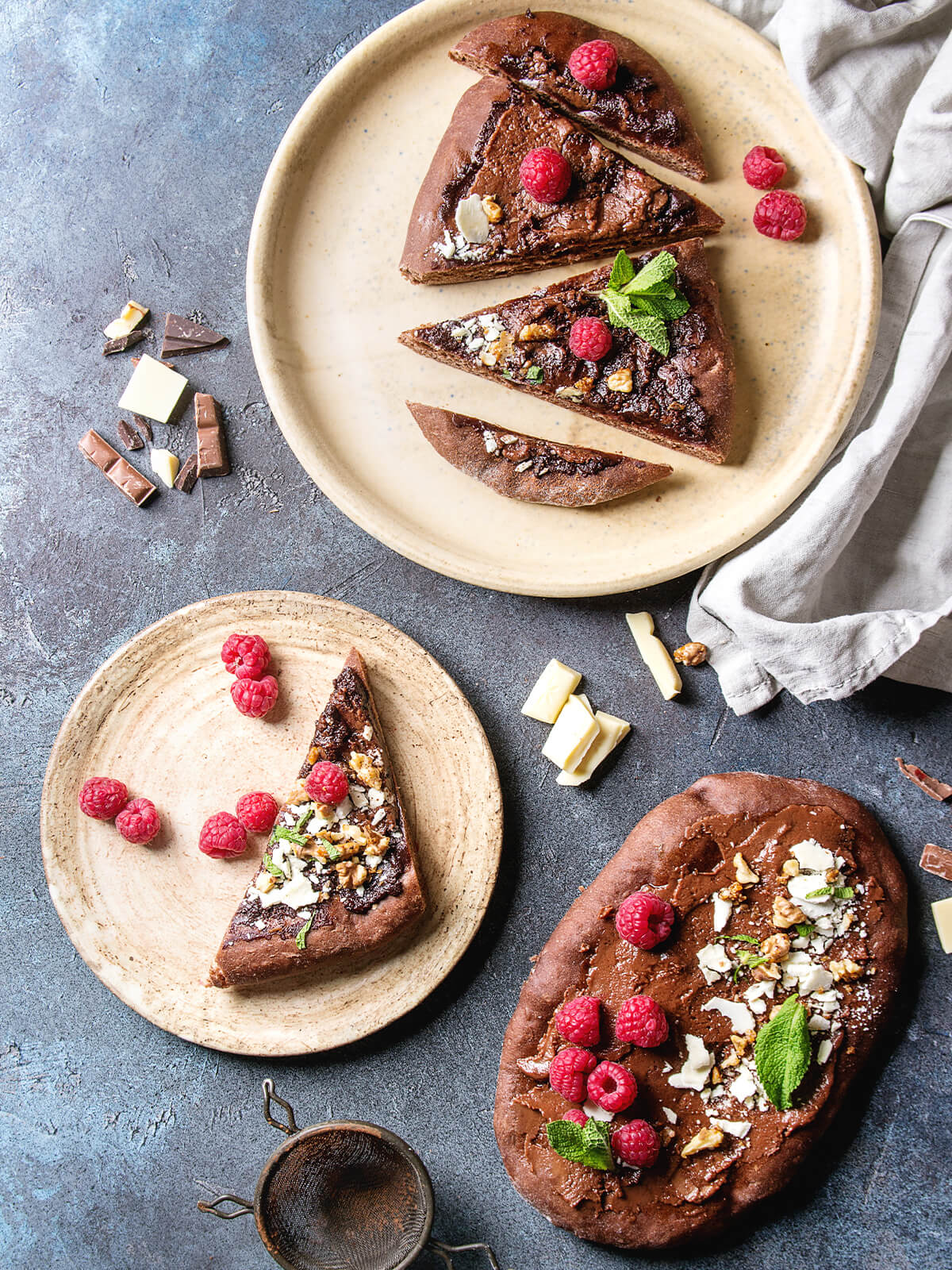 Stücke der ovalen Schoko-Pizza auf hellen Tellern, daneben ein ganzer belegter Kakao-Fladen mit Himbeeren und weißer Schokolade