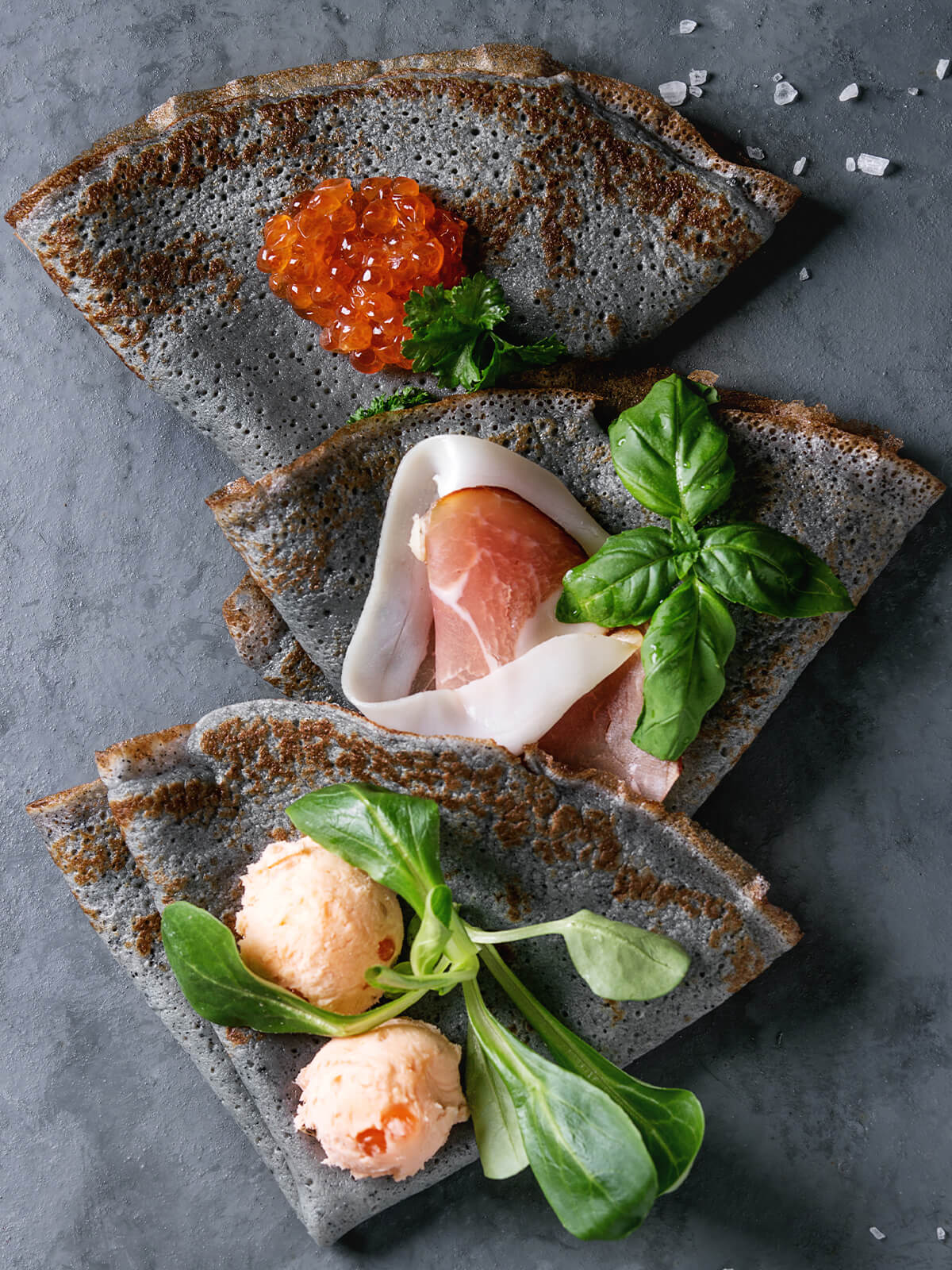Drei schwarze, zu Dreiecken gefaltete Crêpes mit Lachscreme-Nocken, Feldsalat, rotem Kaviar, krauser Petersilie, Rohschinken und Basilikum