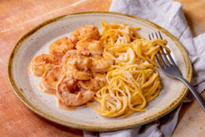 Angerichteter Teller mit Garnelen in heller Tomaten-Sahne-Sauce neben goldgelben Eier-Linguine mit Parmesan, daneben eine Gabel auf grauer Leinenserviette