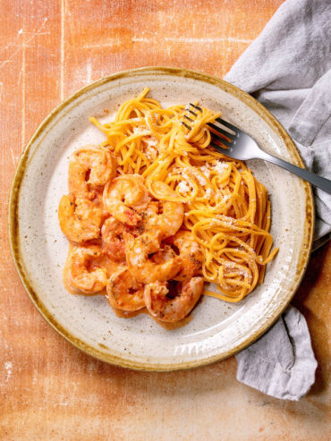 Goldgelbe Eier-Linguine mit fein geriebenem Parmesan, daneben Garnelen in heller Tomaten-Sahne-Sauce; Gabel und graue Leinenserviette liegen am Tellerrand