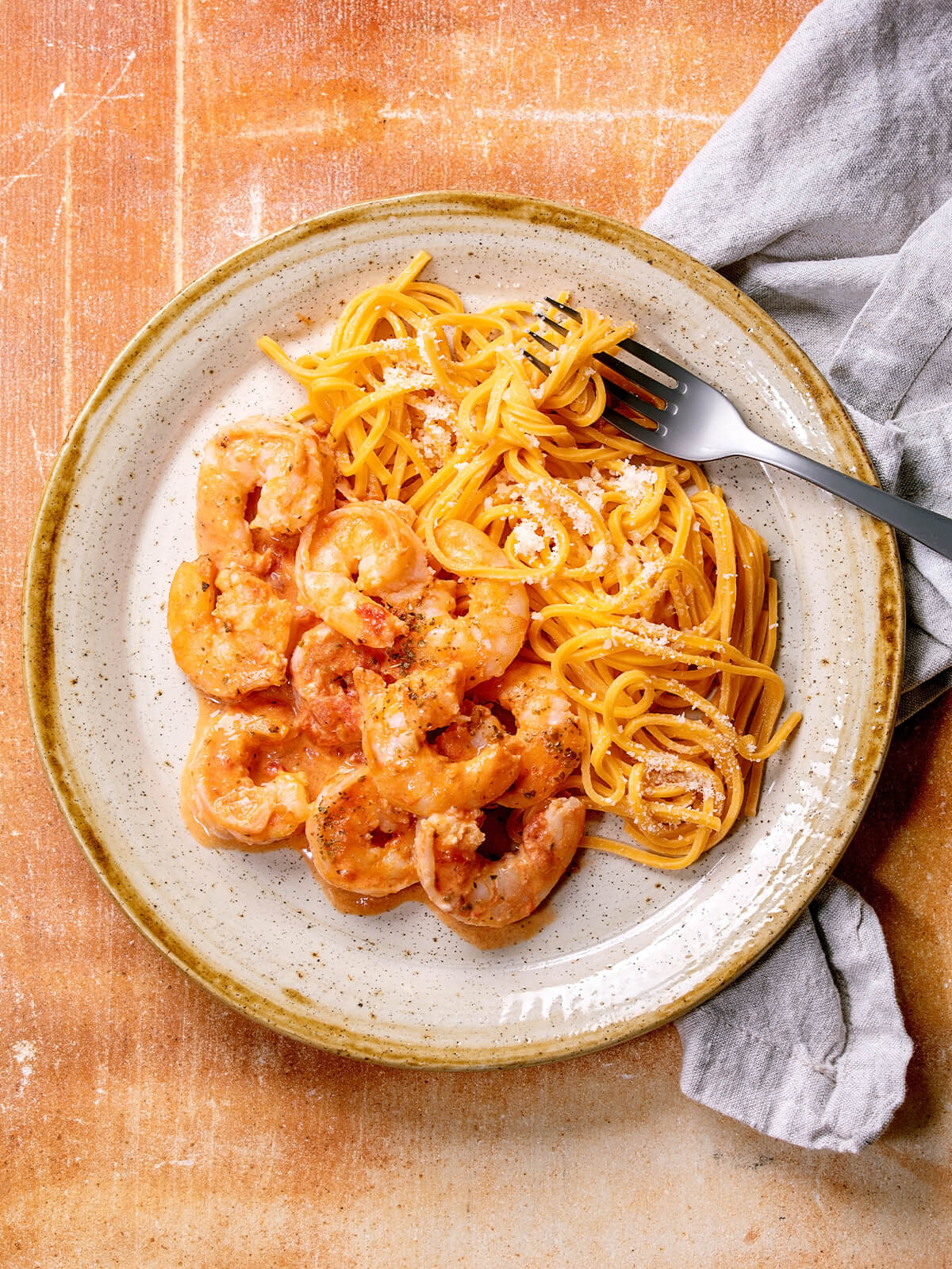 Goldgelbe Eier-Linguine mit fein geriebenem Parmesan, daneben Garnelen in heller Tomaten-Sahne-Sauce; Gabel und graue Leinenserviette liegen am Tellerrand