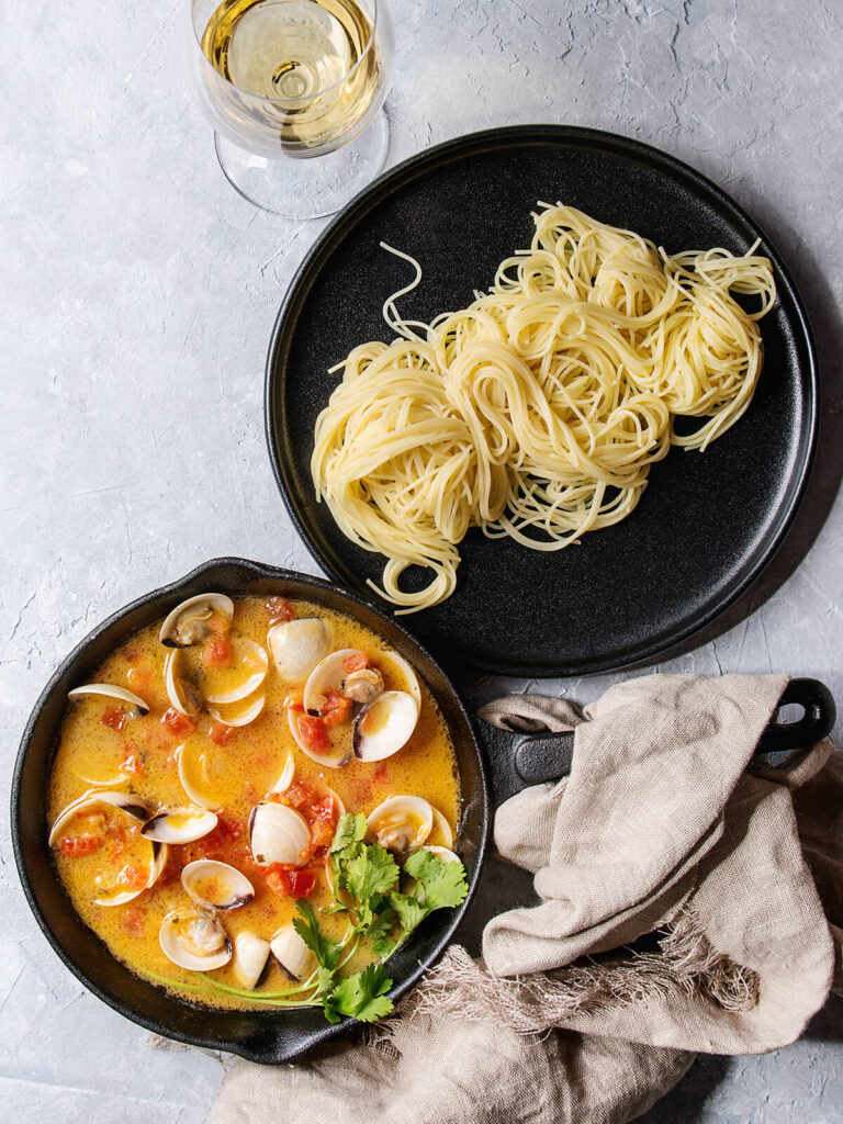 Gusseisen-Pfanne mit Vongole-Sauce neben einem Teller Spaghetti und einem Glas Weißwein