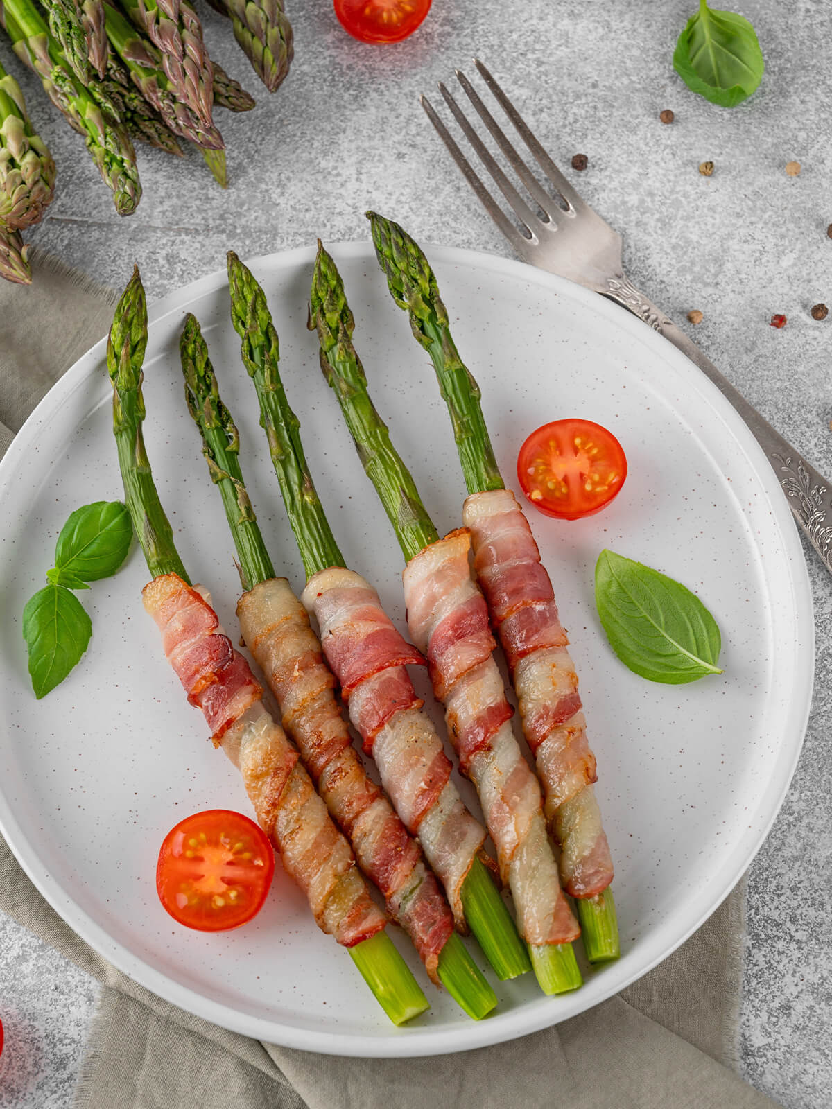 Fünf bacon-umwickelte grüne Spargelstangen auf weißem Teller mit Cherrytomaten