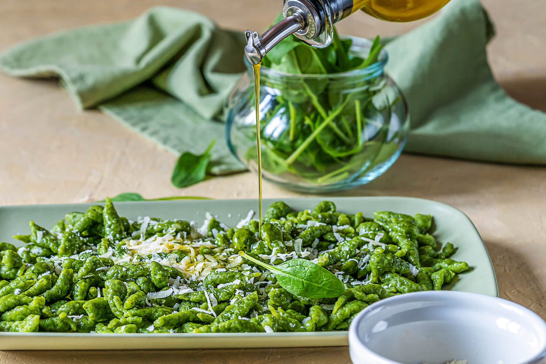 Olivenöl wird aus einer Karaffe über eine Platte mit grünen Spinat-Spätzle und Parmesan geträufelt